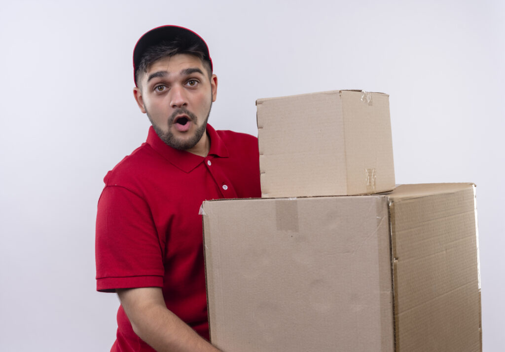 young delivery man in red uniform and cap holding large cardboard boxes looking surprised and amazed standing over white background